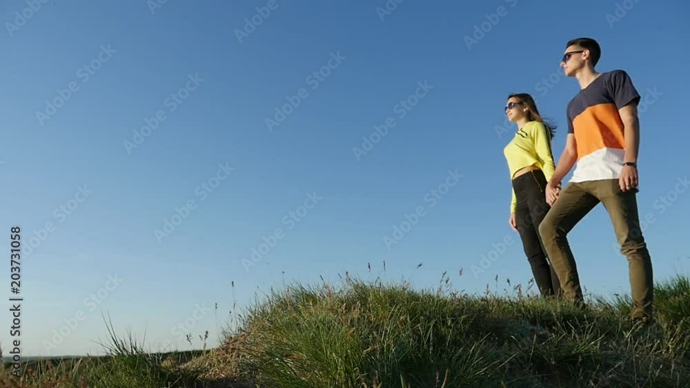 Profile of a sportive young man in sunglasses keeping the hand of his sweetheart with long loose hair on a hilly seashore. They are in love Profile of a sportive young man in sunglasses keeping the hand of his sweetheart with long loose hair on a hilly seashore. They are in love