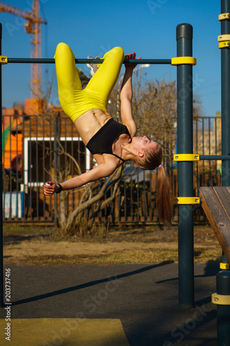 Girl on street workout. She trains press on bar. Girl is hanging upside down. She is dressed in black topic and yellow sports pants.