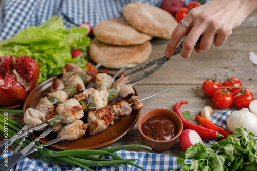 The hand holding the forged fork tends to juicy kebabs. Meat cooked on an open fire. Still-life on a wooden background. Close-up.
