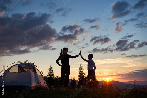Silhouette of a young woman and a child giving each other a high five near a camping at dawn on top of a mountain. A beautiful landscape of fir-trees and sun rising over the mountains and hills © anatoliy_gleb