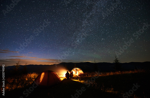 Rear view of silhouette mother and two sons hikers at camping in mountains  sitting on log beside campfire  two tents  looking at amazing night sky full of stars and Milky way  enjoying evening scene