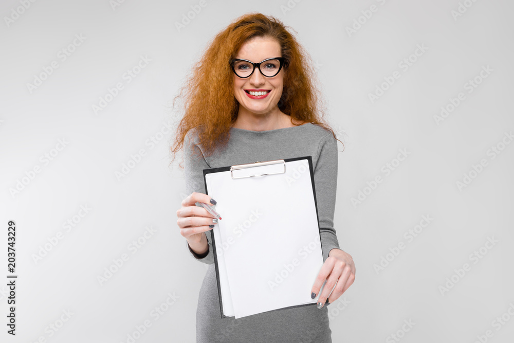 Naklejka premium Charming young girl with red hair. A young girl in a gray sweater. Young girl on a gray background. A young girl with glasses holds a pen and a notebook in her hands