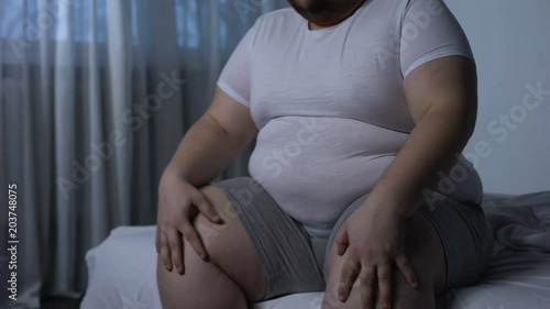 Fat male massaging knees sitting on bed, joint inflammation, orthopedic disorder photo