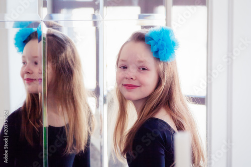A girl with a blue bow in her hair stands near the mirror and looks at the camera and smiles