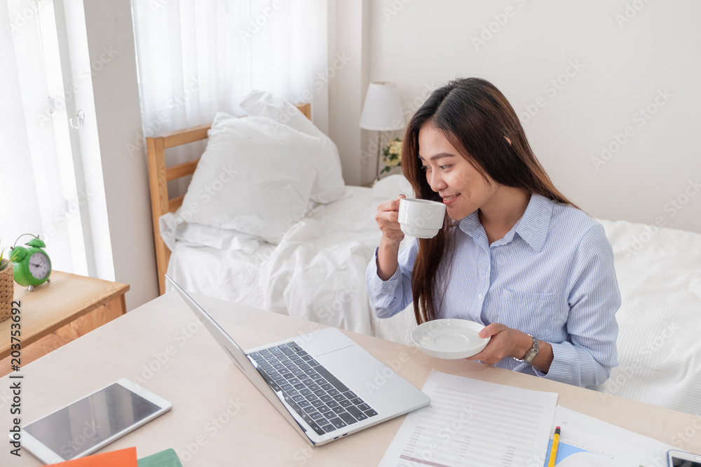 Asian businesswoman drinking coffee and while working with laptop on table in bedroom at home.Work at home concept.work from home concept. Asian businesswoman drinking coffee and while working with laptop on table in bedroom at home.Work at home concept.work from home concept.