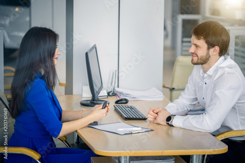 Side view of dialogue between woman customer and car dealership worker sitting at desk, at dealership office photo