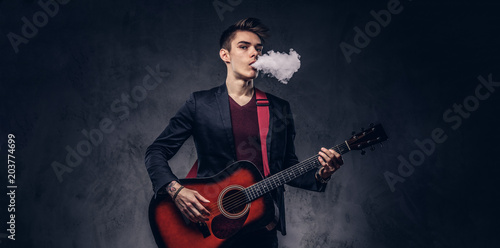 Handsome young musician with stylish hair in elegant clothes exhales smoke while playing acoustic guitar.