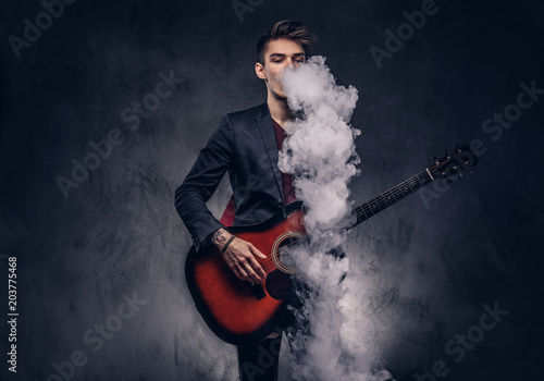 Handsome young musician with stylish hair in elegant clothes exhales smoke while playing acoustic guitar.