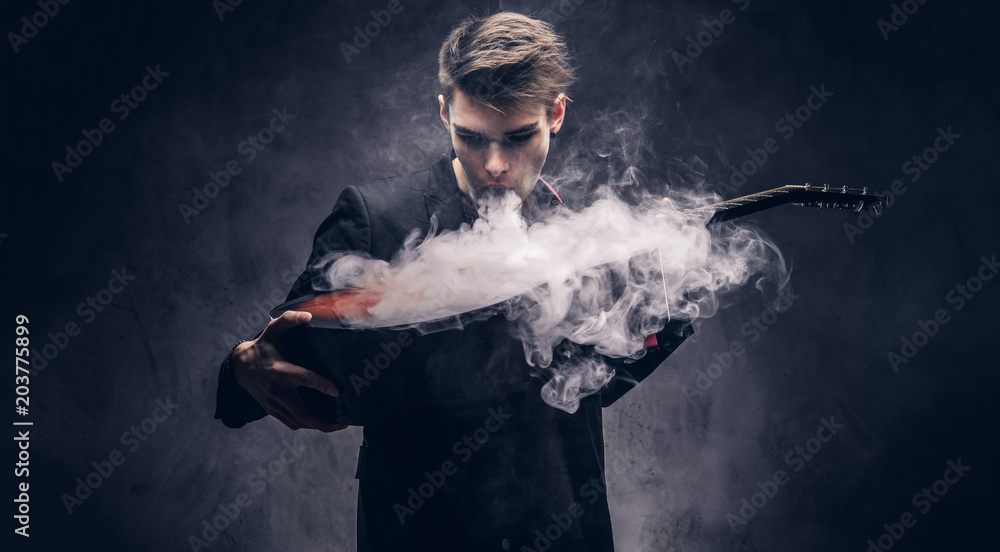 Obraz premium Handsome young musician with stylish hair in elegant clothes exhales smoke on his acoustic guitar.
