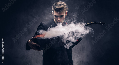 Handsome young musician with stylish hair in elegant clothes exhales smoke on his acoustic guitar.