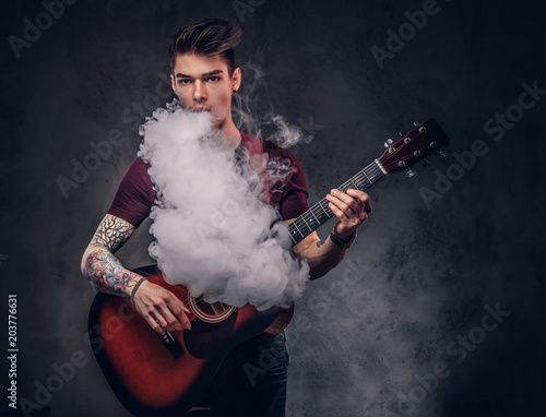Handsome young musician with stylish hair in a t-shirt, exhales smoke while playing acoustic guitar.