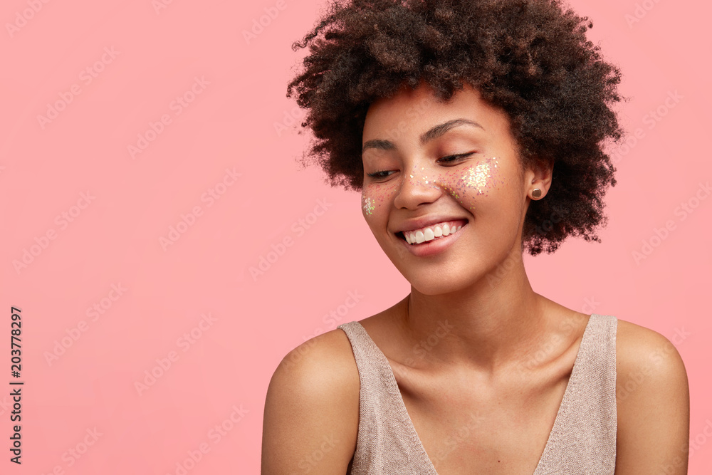 Close up shot of pleasant looking smiling young female with Afro hairstyle and art make up, looks joyfully down, isolated over pink background with copy space for advertisement. Dark skinned woman Close up shot of pleasant looking smiling young female with Afro hairstyle and art make up, looks joyfully down, isolated over pink background with copy space for advertisement. Dark skinned woman