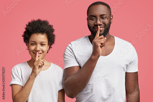 Photo of pleased African American boy and adult, keep fore fingers on lips, demonstrate silence sign and ask to be quiet, pose together on pink studio background. Hush, don`t tell private information photo