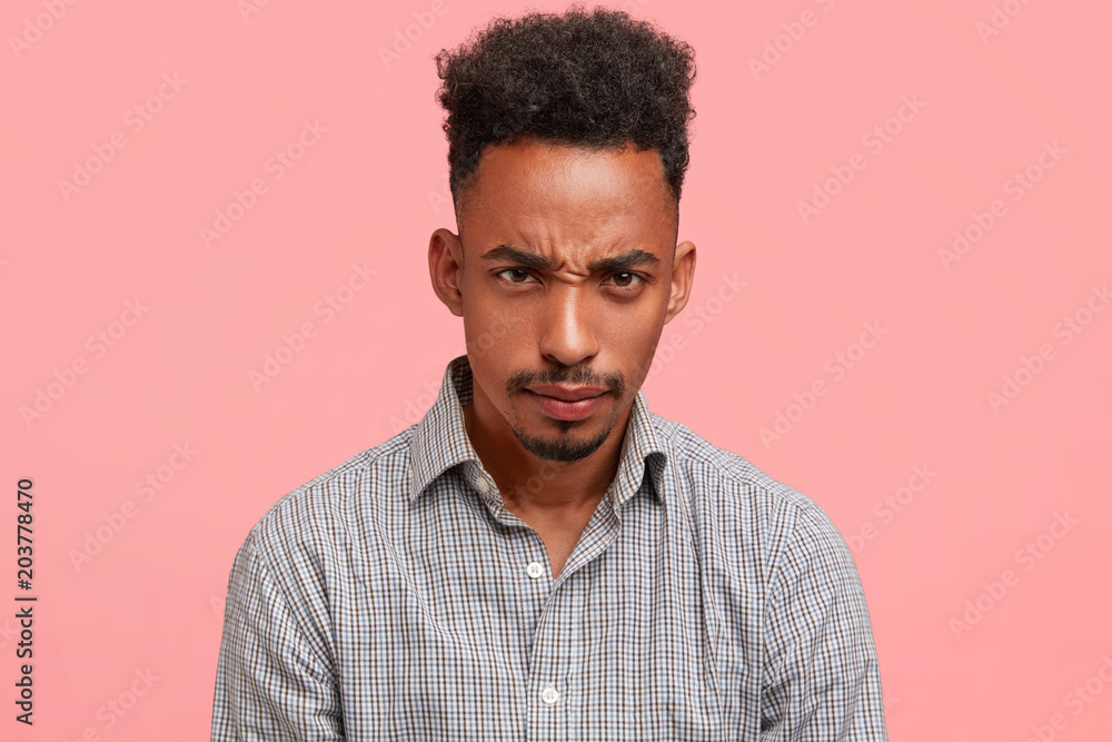 Negative facial expressions concept. Indoor shot of sullen grumpy African American male with trendy haircut, looks with displeased expression, being annoyed and unhappy, has much work on weekends Negative facial expressions concept. Indoor shot of sullen grumpy African American male with trendy haircut, looks with displeased expression, being annoyed and unhappy, has much work on weekends