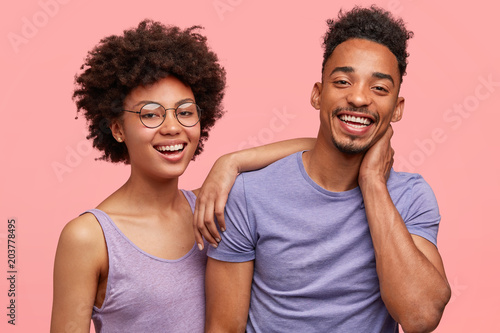Positive African American couple have fun together, smile positively, embrace and support each other, pose together against pink background. Joyful dark skinned woman and her best dark skinned friend