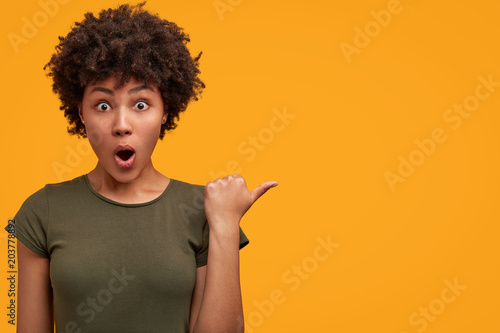 Photo of greatly surprised dark skinned female with bushy Afro haircut, wears casual t shirt, indicates at yellow blank copy space, keeps mouth widely opened, being astonished with something. photo