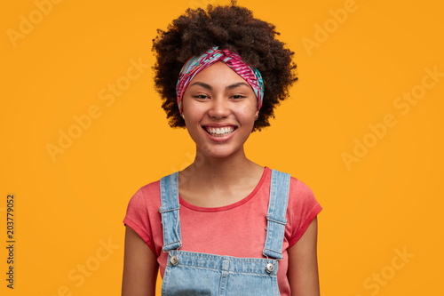 Fashionable dark skinned female teenager with broad smile, has white teeth, satisfied after walk with boyfriend, wears stylish denim overalls, poses against yellow background. Positiveness concept © Wayhome Studio