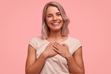 Isolated shot of delighted beautiful female with pleasant smile, hears heart piercing story, keeps hands on chest, wears brackets, isolated over pink studio backgound. Happy Caucasian woman.