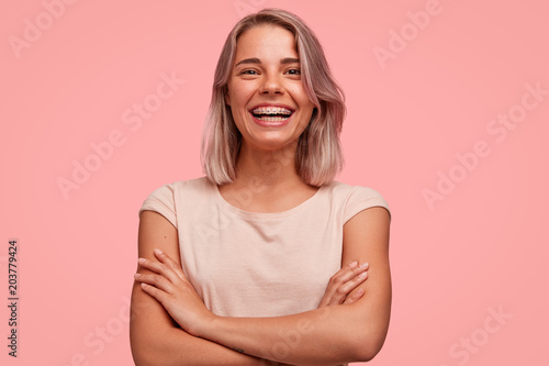 People, happiness and facial expressions concept. Pretty young woman with broad shining smile, keeps hands crossed, being in high spirit, wears braces on teeth, poses alone against pink background photo