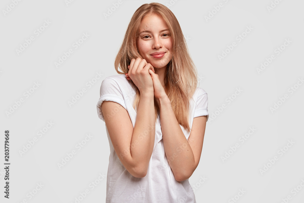 Portrait of delighted young Caucasian female with positive expression, keeps hands together, has gentle smile, being glad to hear good news, poses against white concete wall in casual clothing Portrait of delighted young Caucasian female with positive expression, keeps hands together, has gentle smile, being glad to hear good news, poses against white concete wall in casual clothing