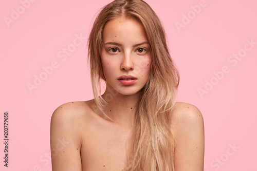 Close up shot of pleasant looking European female with confident expression, looks seriously directly at camera, poses nude against pink background, demonstrates her beauty and healthy skin.