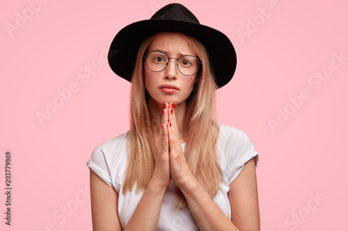 Horizontal shot of pretty female student pleads for good mark, looks with pleading expression, keeps palms pressed together, asks for help, poses against pink background. Please, forgive me!