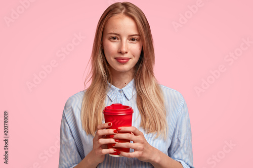 Young cute female office worker with lovely appearance, has coffee break, holds red paper cup of takeaway coffee, poses against pink studio background. People, leisure and relaxation concept