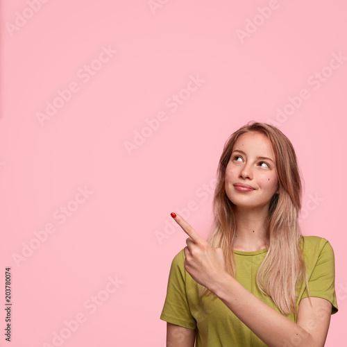 Vertical shot of good looking female model advertises new product in studio, points at blank copy space, isolated over pink background. Pretty woman seller attracts your attention to something
