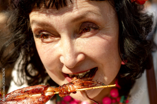 Adult woman is eating squid with skewers in the summer outdoors. Portrait.