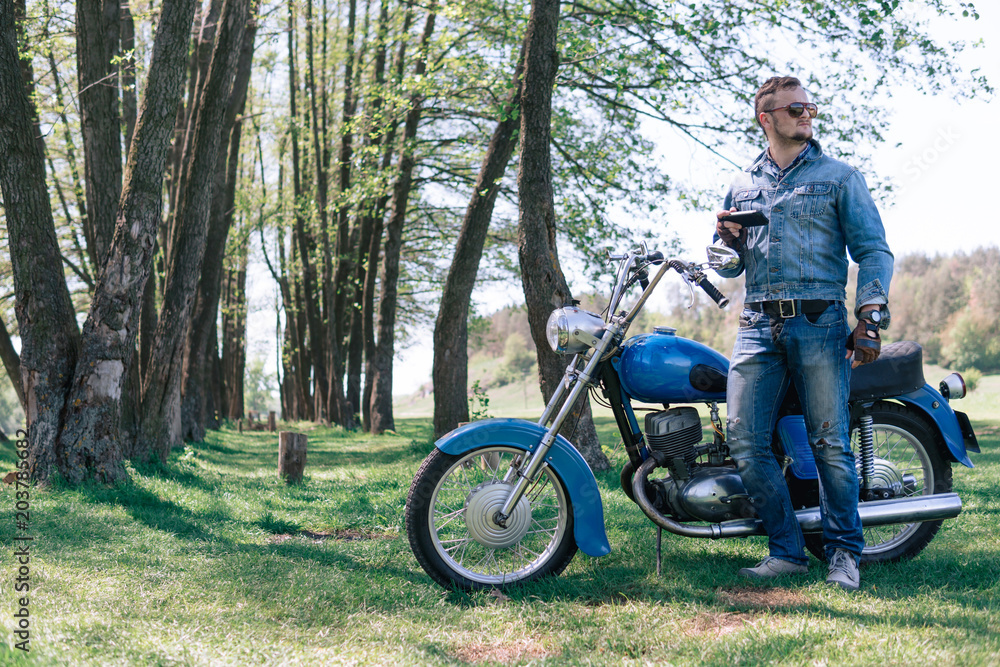 Portrait young guy with sunglasses and Jeans jacket and pants posing sitting on his vintage retro motorbike. biker wearing jeans, fashion men. old timer age concept, 1960s style, outdoor dirt road