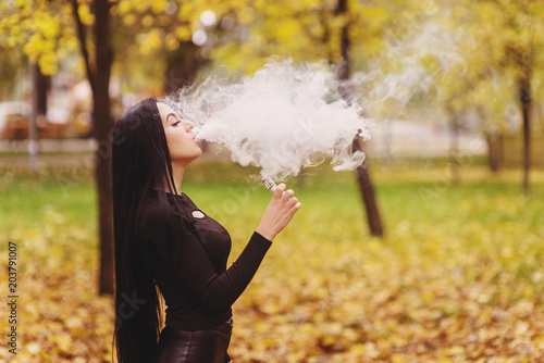 Vaping. Young woman in black clothes, vaping device at the autumn park. Toned image photo