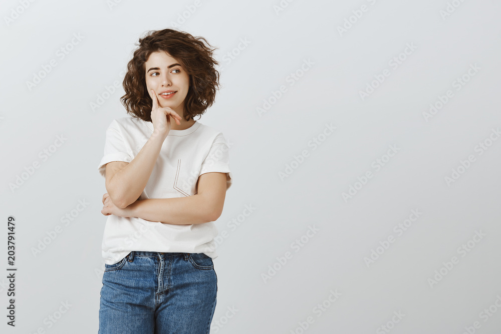 Smoothie or juice, hard choice. Portrait of attractive caucasian curly-haired girl looking aside and holding fingers on cheek, thinking, having idea while working on project over gray background Smoothie or juice, hard choice. Portrait of attractive caucasian curly-haired girl looking aside and holding fingers on cheek, thinking, having idea while working on project over gray background