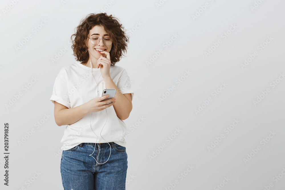 Worrying girl hesitates to send message with confession in love. Anxious attractive curly-haired caucasian woman, smiling nervously, touching lip, listening music in earphones and holding smartphone Worrying girl hesitates to send message with confession in love. Anxious attractive curly-haired caucasian woman, smiling nervously, touching lip, listening music in earphones and holding smartphone