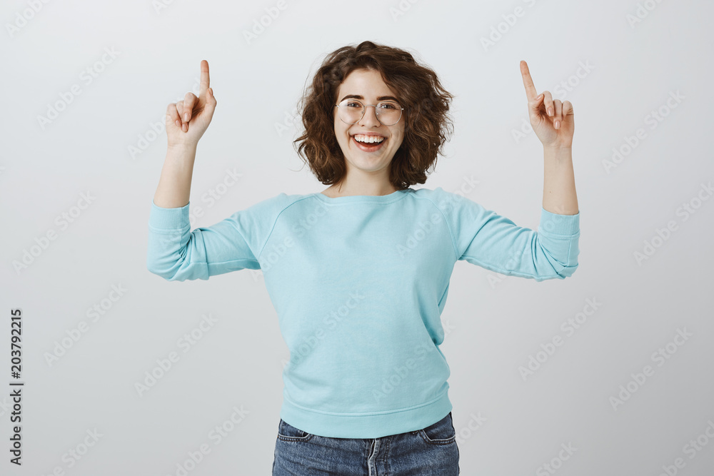 Attractive museum guide showing tourists around. Portrait of happy joyful female student in glasses, feeling excited and carefree, raising index fingers and pointing upwards over gray wall Attractive museum guide showing tourists around. Portrait of happy joyful female student in glasses, feeling excited and carefree, raising index fingers and pointing upwards over gray wall