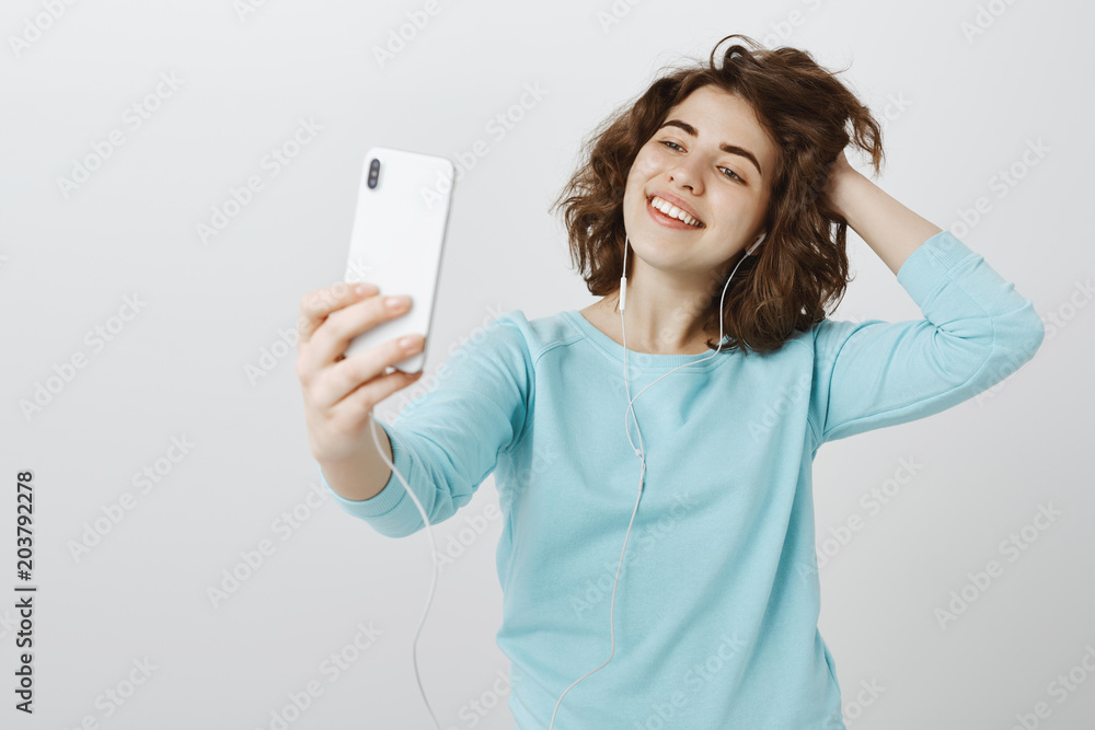 Fototapeta premium Time for selfie. Portrait of stylish confident good-looking female student in earphones, pulling hand with smartphone, taking photo and touching hair playfully, being happy over gray wall