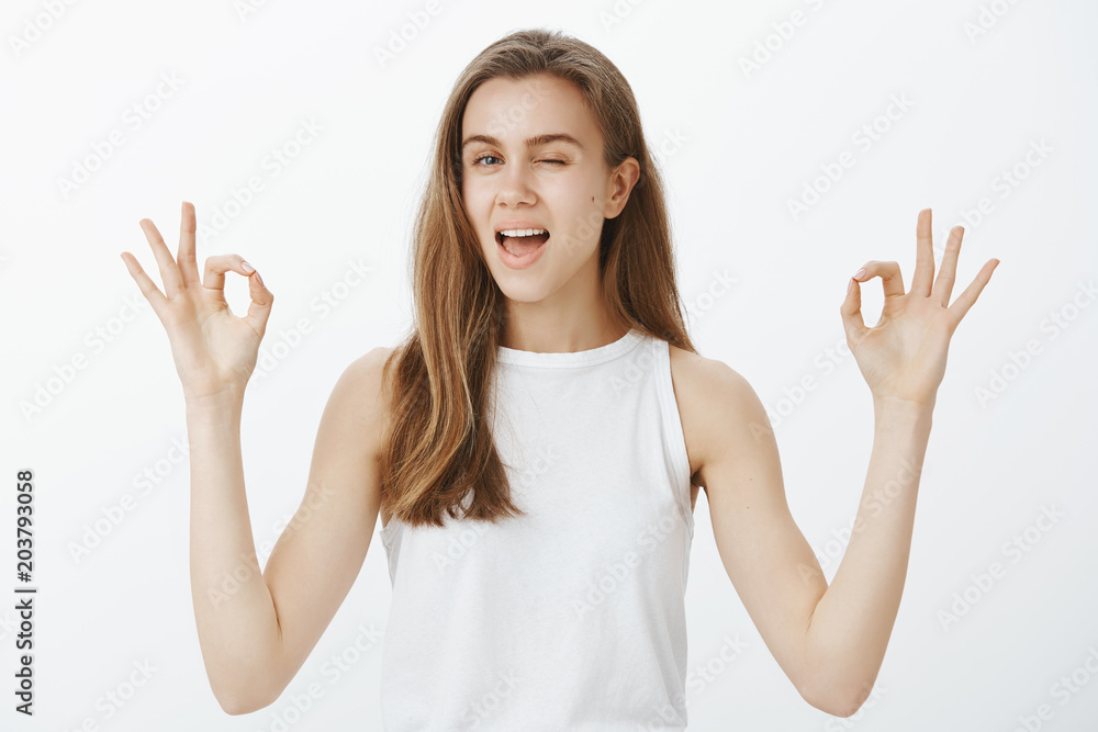 No worried and problems. Confident friendly-looking attractive woman with blond hair in white top, raising okay sign, winking with broad smile, standing over gray wall, giving approval or saying ok No worried and problems. Confident friendly-looking attractive woman with blond hair in white top, raising okay sign, winking with broad smile, standing over gray wall, giving approval or saying ok