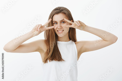 Waist-up shot of positive charming female employee with fair hair, showing victory or peace gestures over eyes and smiling happilly, feeling boost of joy while dancing disco over gray background