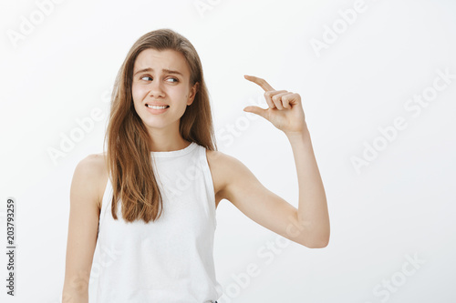 Waist-up shot of disappointed awkward attractive woman in white tank-top, raising hand and shaping small item, looking at fingers and frowning from dislike and regret, standing over gray background