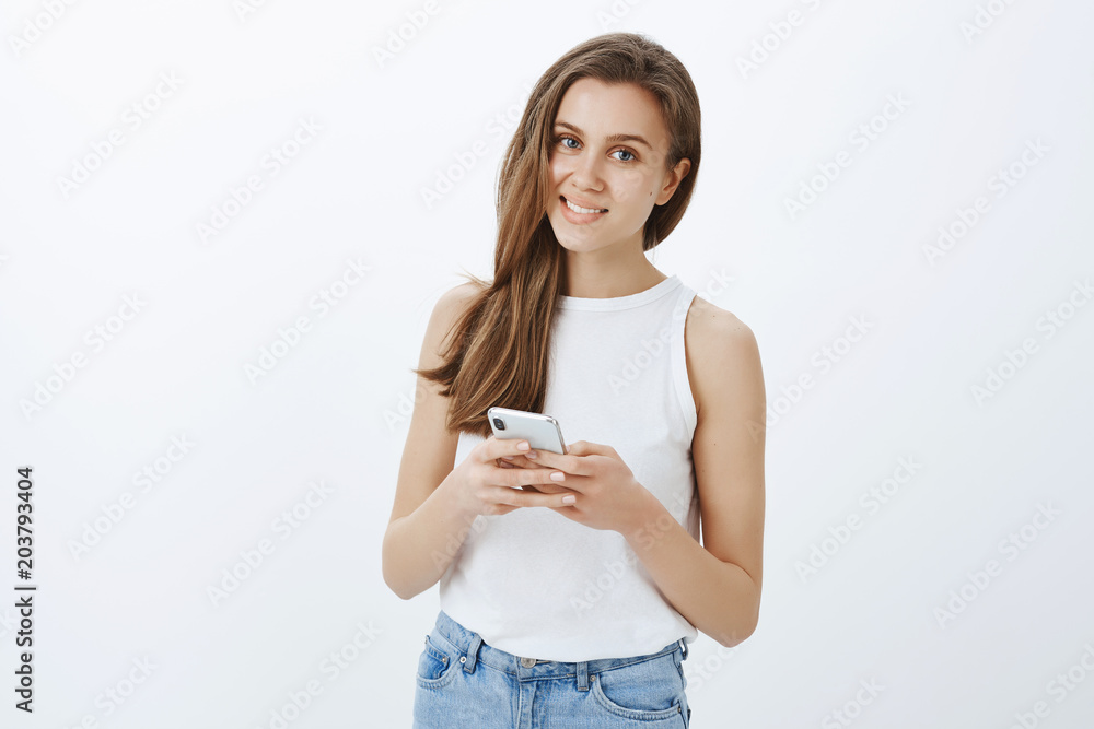Indoor shot of good-looking carefree female with confident expression, holding smartphone and gazing with smile at camera, being intrigued by question while typing message or making notes in device Indoor shot of good-looking carefree female with confident expression, holding smartphone and gazing with smile at camera, being intrigued by question while typing message or making notes in device
