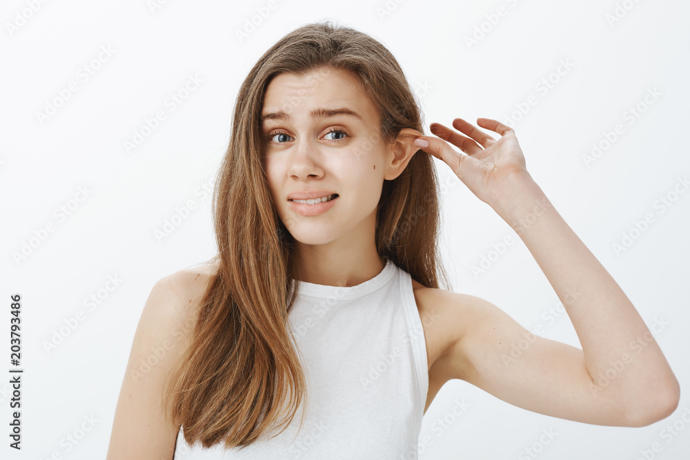 Stop lying my ears tired. Portrait of funny attractive woman with fair hair,  pulling ear with hand and staring displeased at camera, feeling awkward  while standing against gray background Stock Photo |