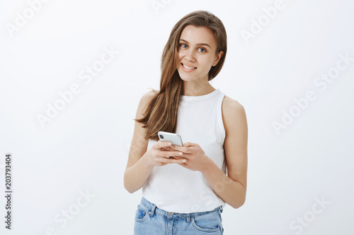 Indoor shot of good-looking carefree female with confident expression, holding smartphone and gazing with smile at camera, being intrigued by question while typing message or making notes in device