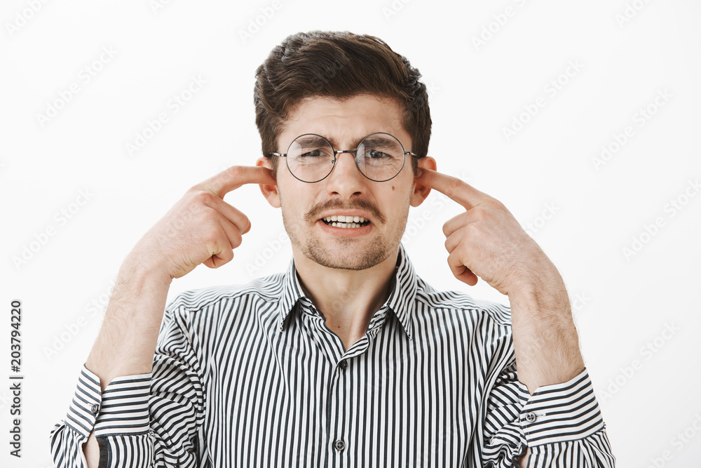 Portrait of upset whining ordinary guy in round glasses and striped shirt, covering ears with index fingers, making displeased expression, feeling dislike while hearing awful scratch on blackboard