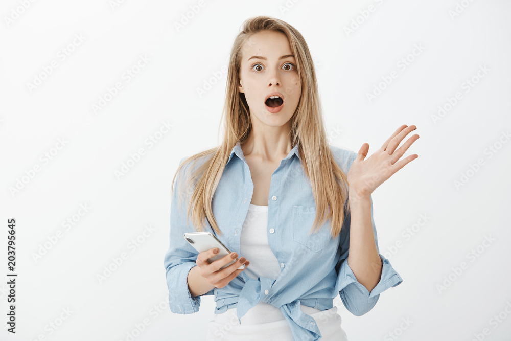 Indoor shot of impressed amazed attractive fair-haired woman in stylish outfit, raising palm and dropping jaw while explaining recent news to friend, holding smartphone and gesturing from excitement Indoor shot of impressed amazed attractive fair-haired woman in stylish outfit, raising palm and dropping jaw while explaining recent news to friend, holding smartphone and gesturing from excitement