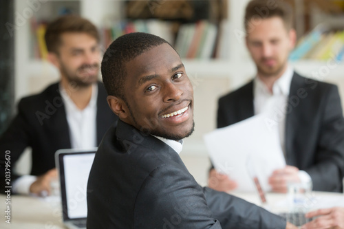 Smiling successful african american looking at camera back over shoulder while sitting at business meeting. Workers discussing company plans and strategies at briefing in background. Diverse team