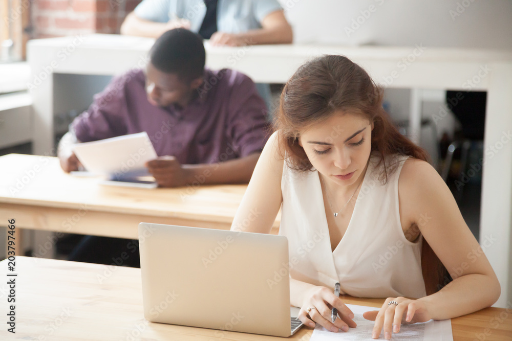 Young female freelancer thoughtfully working with paperwork and laptop, checking report data, evaluating company results in coworking office. Student getting ready for exam, reading handouts Young female freelancer thoughtfully working with paperwork and laptop, checking report data, evaluating company results in coworking office. Student getting ready for exam, reading handouts
