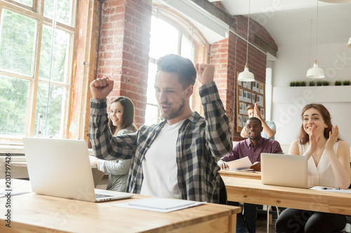 Happy successful worker excited with good results on stock market, raising hands up with yes gesture, colleagues congratulating with achievement. Trader celebrating online win in coworking office.