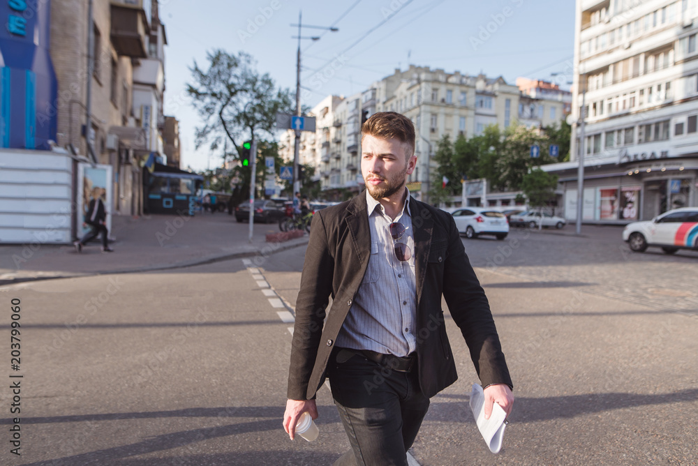 Portrait of a beautiful man in a costume passing a road on a pedestrian crossing. A businessman with documents and a cup of coffee passes the road to the green light of the traffic light. Portrait of a beautiful man in a costume passing a road on a pedestrian crossing. A businessman with documents and a cup of coffee passes the road to the green light of the traffic light.