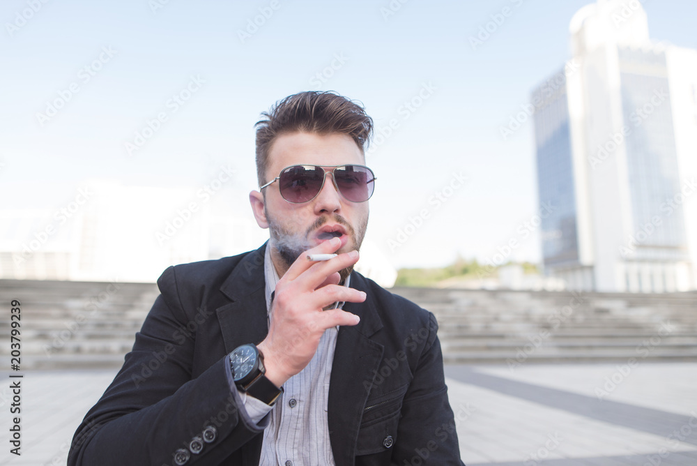 Portrait of a handsome young businessman smiling a cigarette on a city background. A business man smokes a cigarette closeup. Portrait of a handsome young businessman smiling a cigarette on a city background. A business man smokes a cigarette closeup.