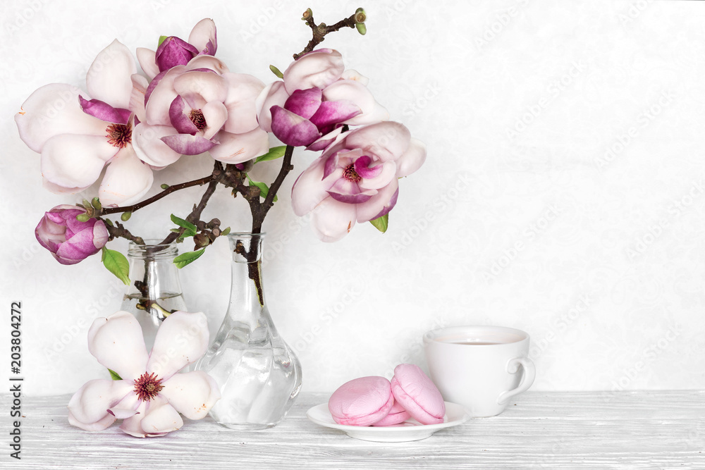 Fototapeta premium Różowa magnolia kwitnie bukiet z filiżanką i macarons na białym drewnianym tle Selekcyjna ostrość