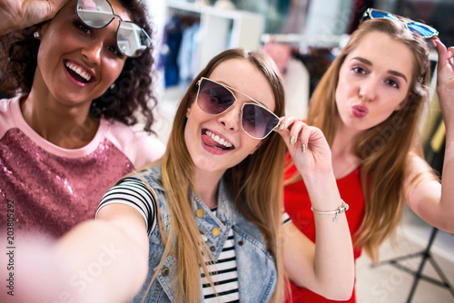 Smiling girlfriends wearing stylish sunglasses having fun time taking selfie with mobile phone while doing shopping in clothing store